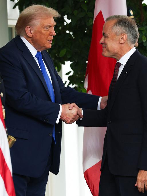 Donald Trump und Mark Carney schütteln einander die Hände. Sie stehen vor der amerikanischen und der kanadischen Flagge.