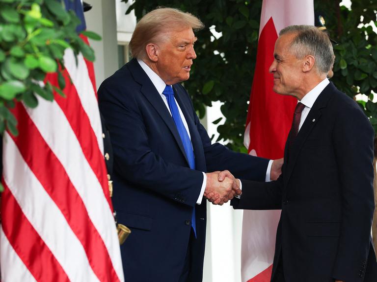 Donald Trump und Mark Carney schütteln einander die Hände. Sie stehen vor der amerikanischen und der kanadischen Flagge.