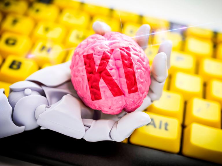 Fotomontage: Roboterhand hält ein Gehirn mit Aufschrift KI vor Computertastatur, Symbolfoto 