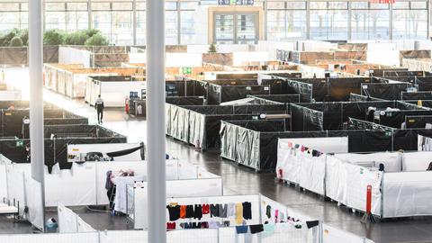 Betten für Geflüchtete stehen mit zum Sichtschhutz aufgestellten Bauzäunen dazwischen in einer Messehalle. Über den Zäunen hängen Kleidungsstücke. 