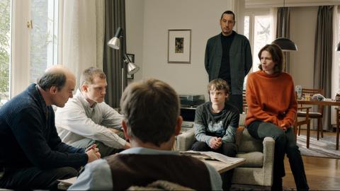 An einem Wohnzimmertisch sitzen Familie und Polizeiermittler beisammen und besprechen die Lage.