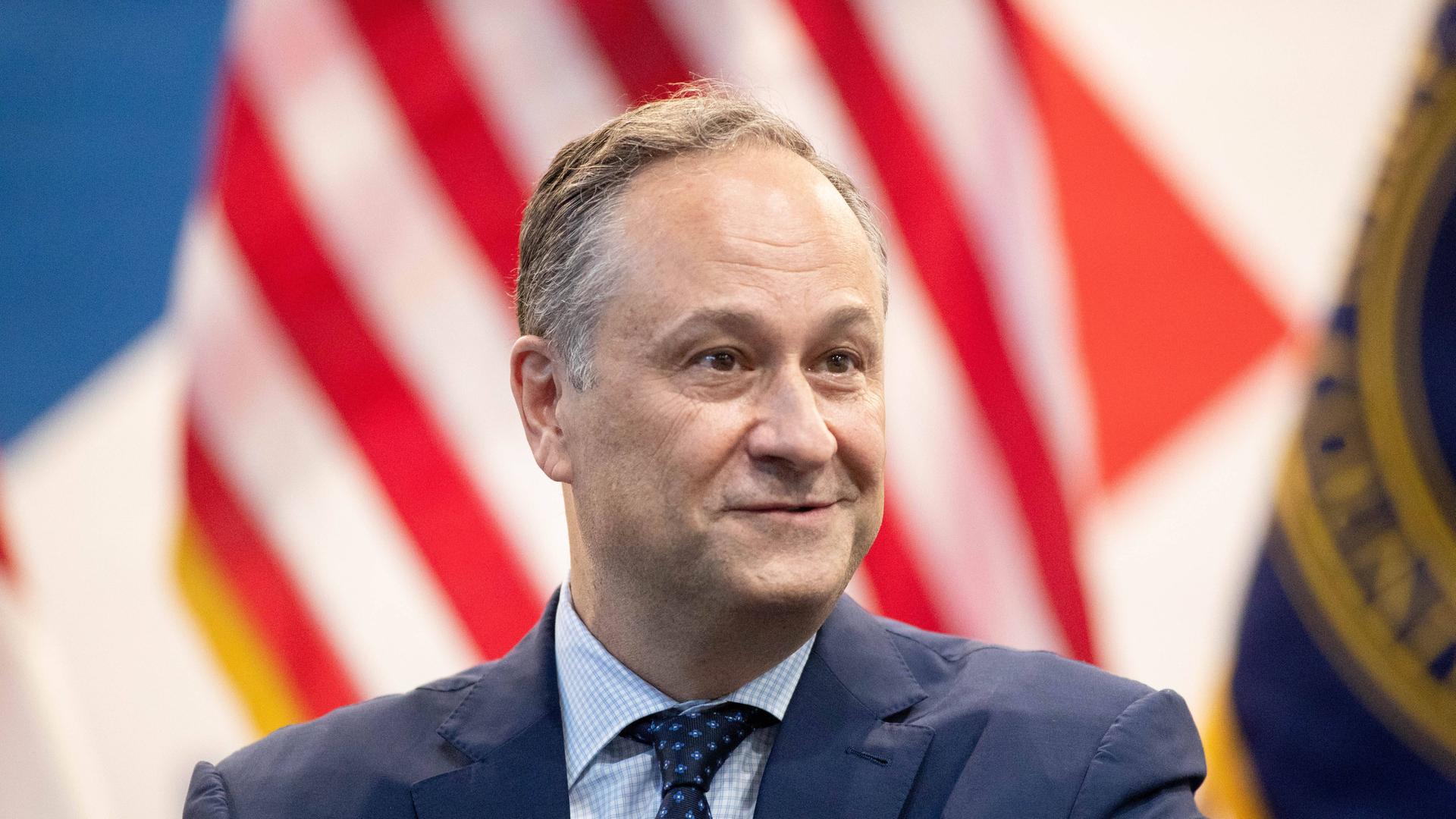 Das Bild zeigt Douglas Emhoff im dunkelblauen Anzug vor einer Flagge der Vereinigten Staaten von Amerika.