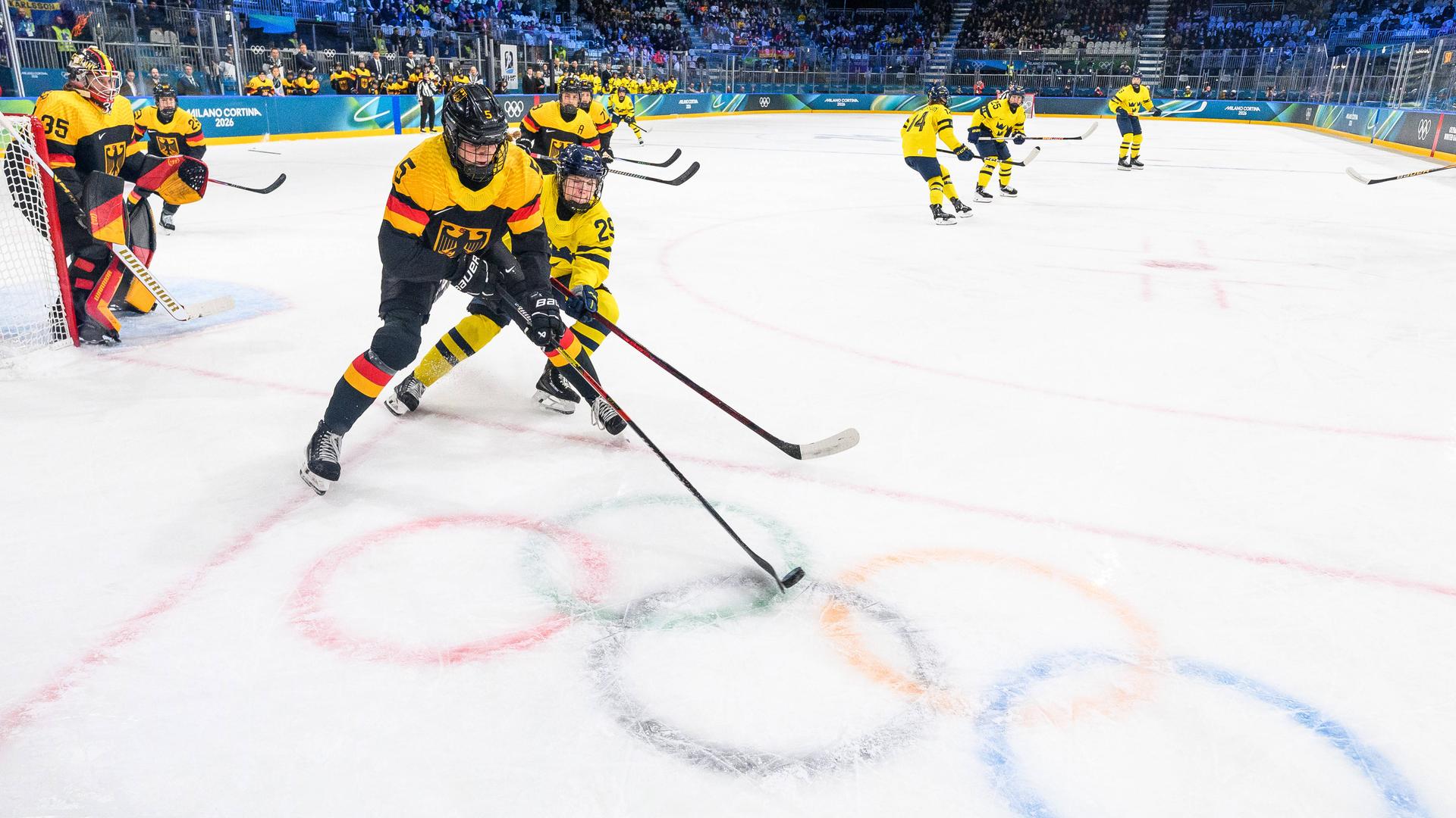 Die deutsche Eishockey-Nationalspielerin Charlott Schaffrath führt den Puck im Duell mit Felizia Wikner Zienkiewicz aus Schweden hinter dem deutschen Tor. Auf dem Eis sind die Olympischen Ringe zu erkennen.