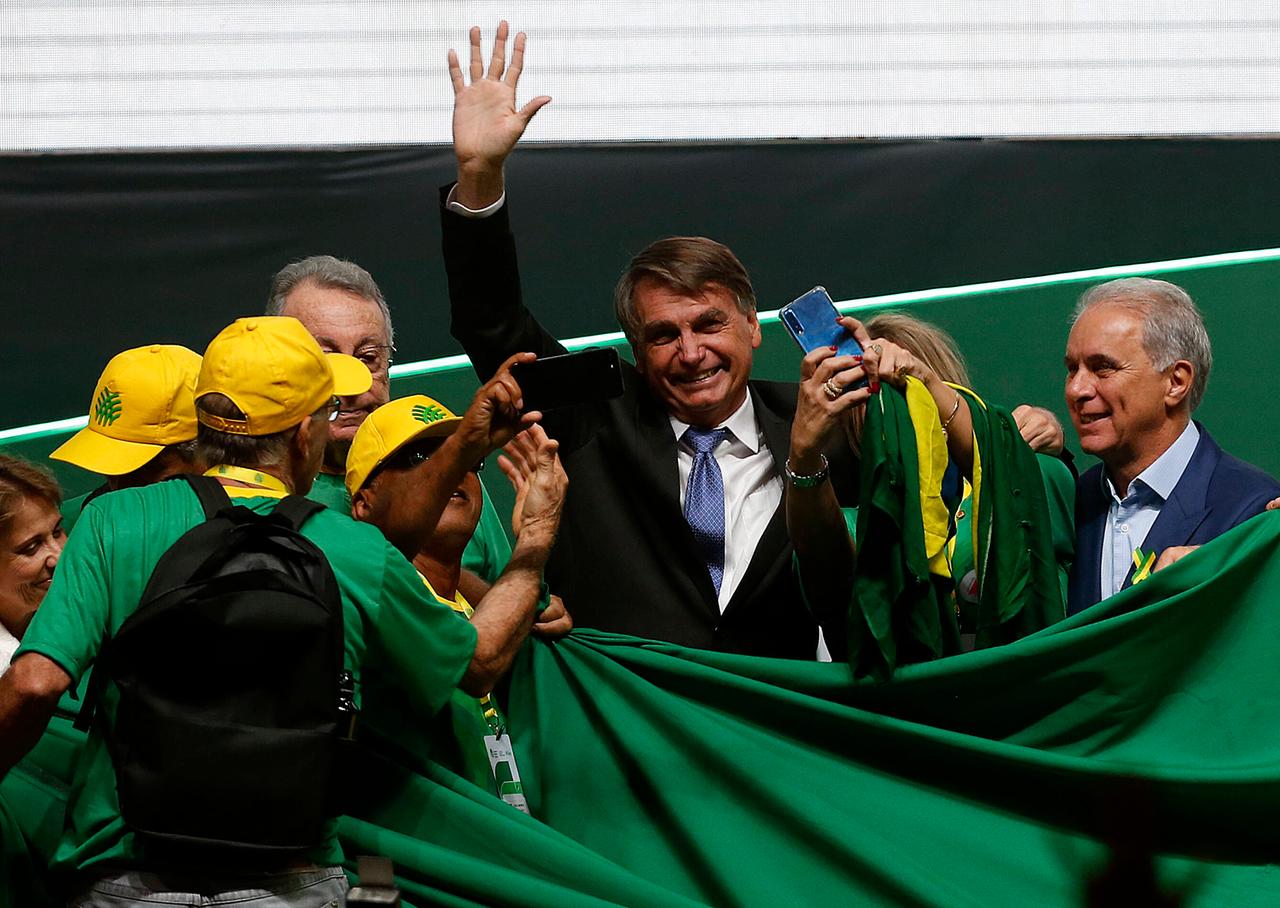 Bolsonaro steht in einer kleinen Schar von Anhängern mit gelben Käppis, überall sind grüne Laken und er winkt ins Publikum.