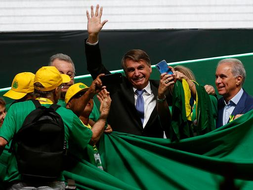 Bolsonaro steht in einer kleinen Schar von Anhängern mit gelben Käppis, überall sind grüne Laken und er winkt ins Publikum.