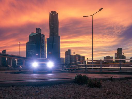 Skyline des Köng Abdullah Finanzdistrikt (KAFD) in Riyadh vor orangenem Himmel. Die Scheinwerfer eines Fahrzeugs leuchten in Richtung der Kamera. Skyline des Köng Abdullah Finanzdistrikt (KAFD) in Riyadh vor orangenem Himmel. Die Scheinwerfer eines Fahrzeugs leuchten in Richtung der Kamera.