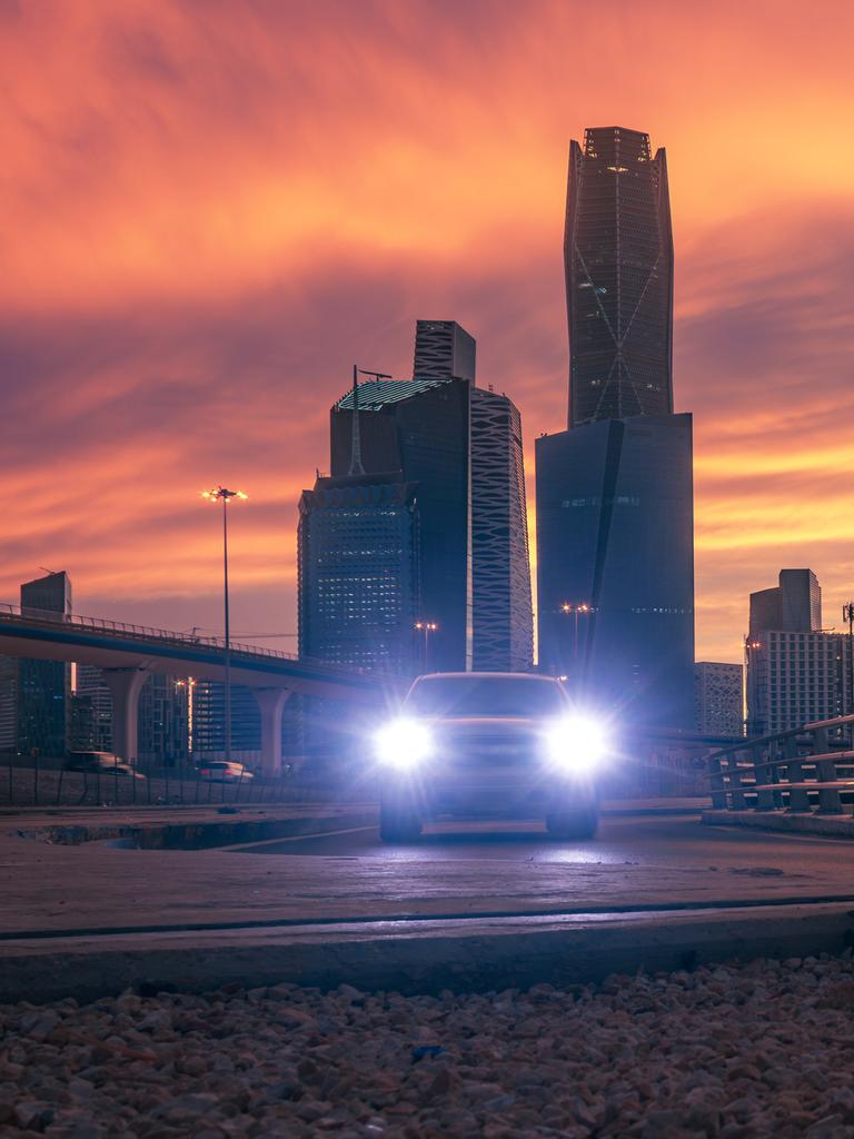 Skyline des Köng Abdullah Finanzdistrikt (KAFD) in Riyadh vor orangenem Himmel. Die Scheinwerfer eines Fahrzeugs leuchten in Richtung der Kamera. Skyline des Köng Abdullah Finanzdistrikt (KAFD) in Riyadh vor orangenem Himmel. Die Scheinwerfer eines Fahrzeugs leuchten in Richtung der Kamera.