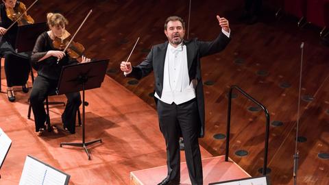 Ein Dirigent im Frack steht auf einem Podest, hält seine Arme erhoben und gibt dem Orchester Zeichen. Vom Orchester kann man nur ein paar Notenständen und Geigerinnen erkennen.
