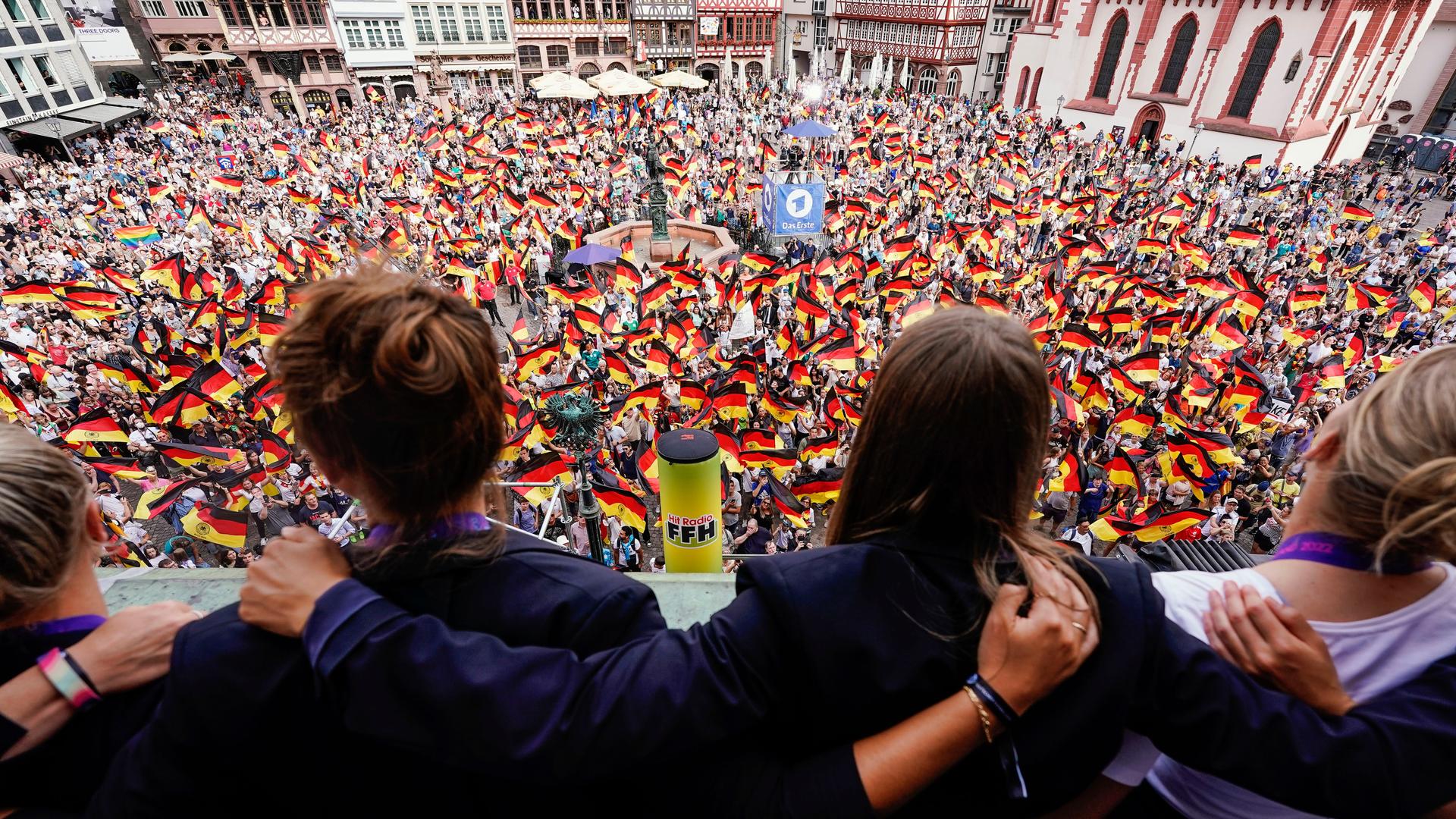 Die Spielerinnen der deutschen Nationalelf schauen von einem Balkon auf eine Masse an feiernden Fans mit Deutschlandfahnen. Mehrere Fernseh- und Radiostationen sind zu erkennen.
