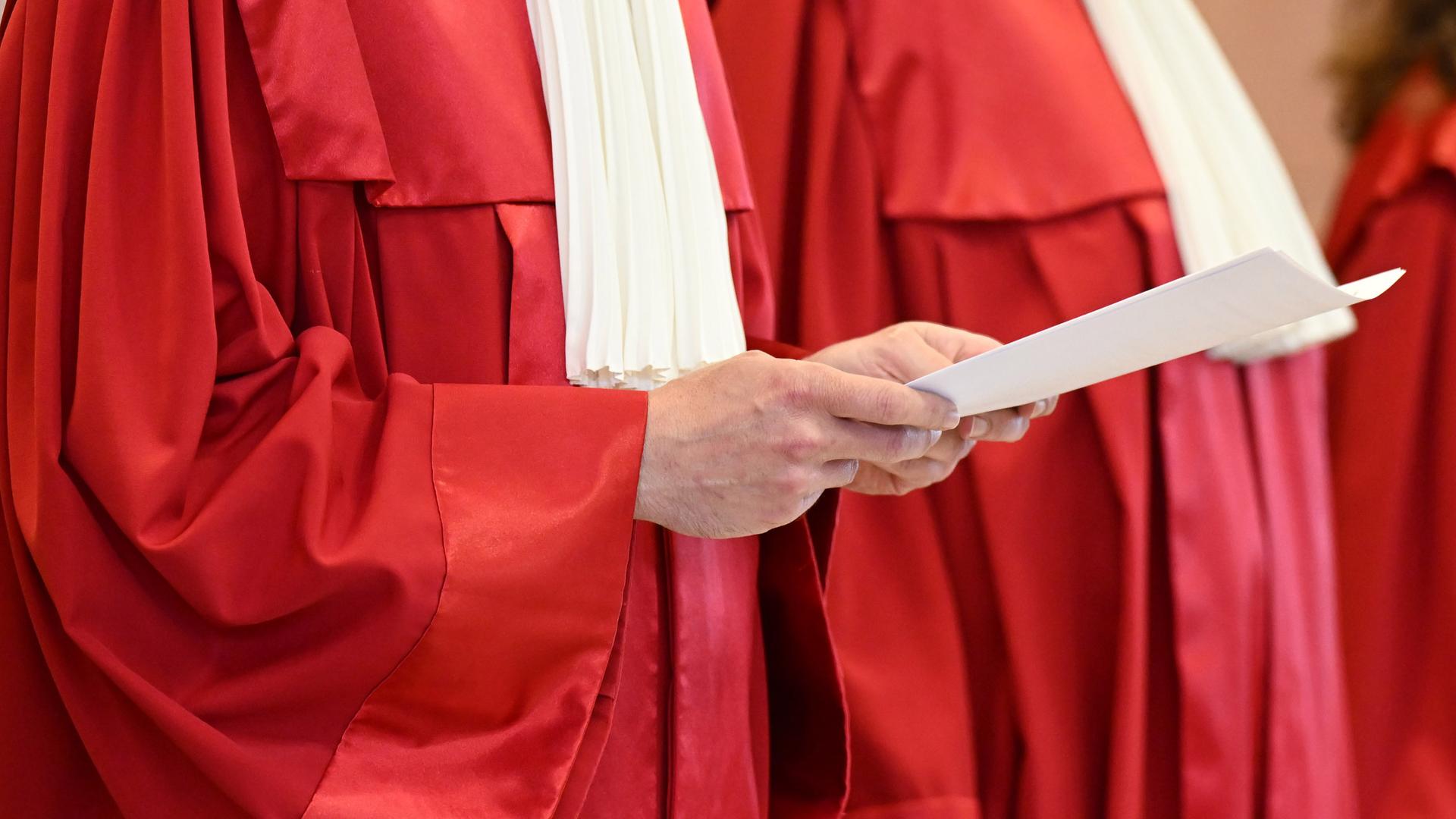 Eine Person in der roten Robe des Bundesverfassungsgerichts hält eine Urteilsverkündung in den Händen.