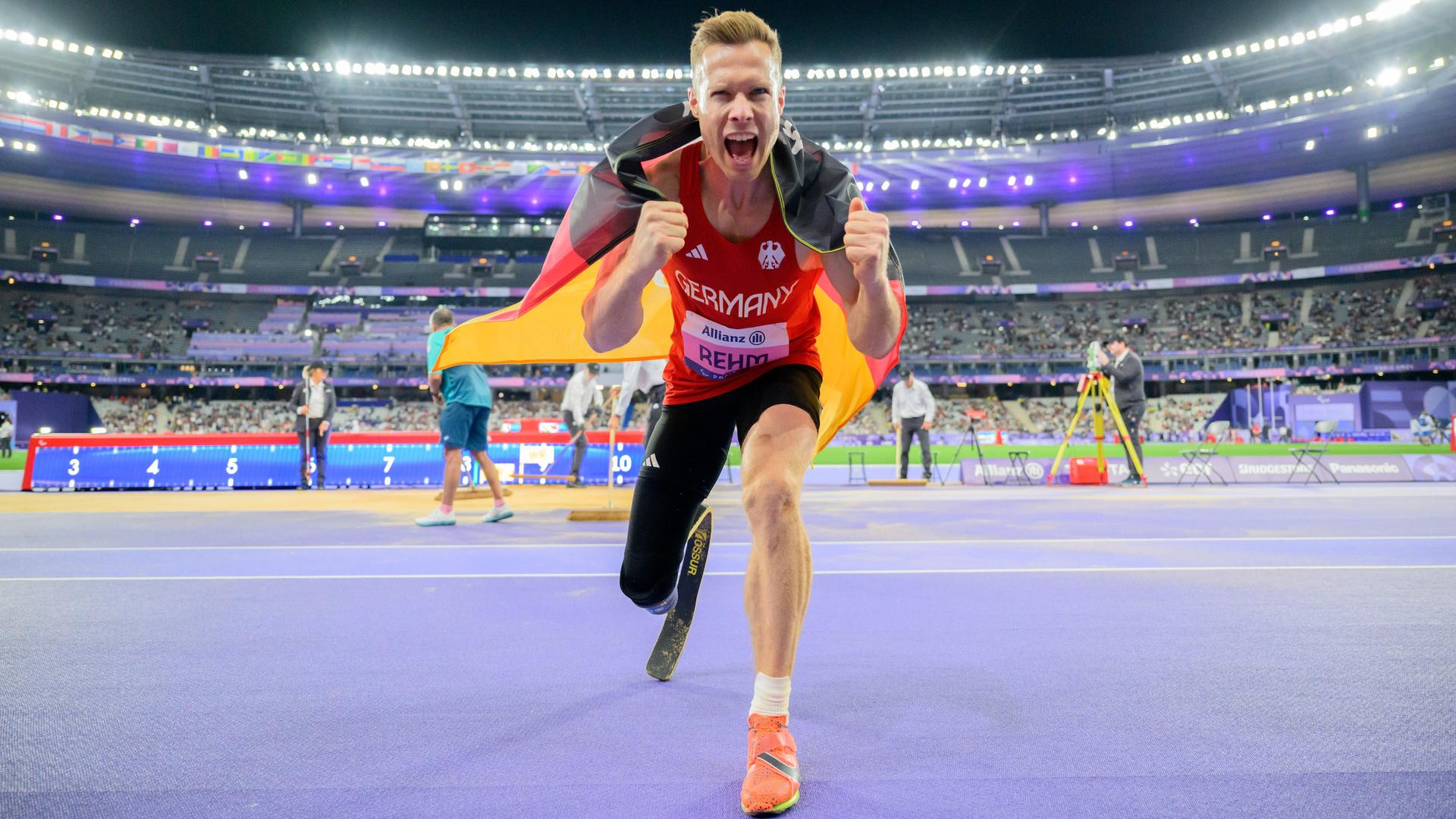 Markus Rehm aus Deutschland feiert seine Goldmedaille. Er hat die Deutschlandfahne um die Schulter gelegt und ballt die Fäuste in Richtung Kamera. Markus Rehm aus Deutschland feiert seine Goldmedaille. Er hat die Deutschlandfahne um die Schulter gelegt und ballt die Fäuste in Richtung Kamera.