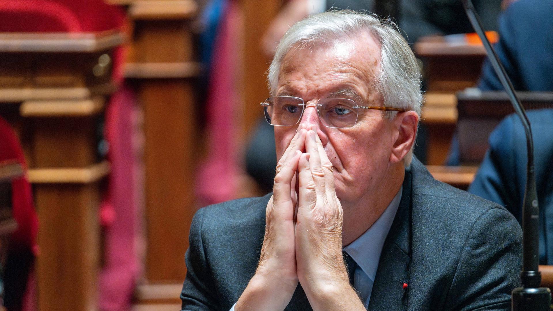 Der neue französische Premierminister Michel Barnier sitzt im Parlament. Er hat die Hände vor dem Gesicht gefaltet. 