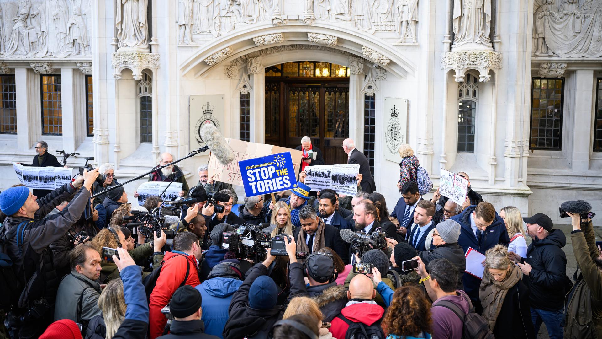 Anwalt Toufique Hossain spricht vor dem Obersten Britischen Gerichtshof mit der Presse. Eine Person in der Menge hält ein Schild mit der Aufschrift "Stop the Tories" in die Höhe.