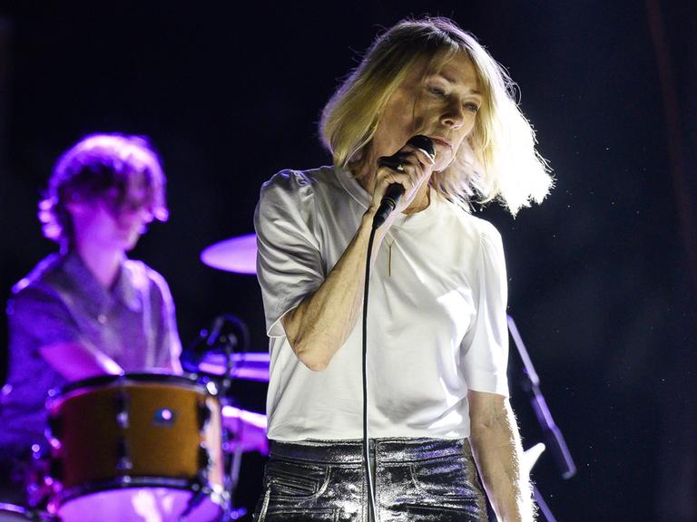 Eine blonde Sängerin - Kim Gordon - steht auf einer Konzertbühne. Sie trägt ein weißes T-Shirt und silbernglänzende Shorts und hält ein Mikrofon in der Hand. Im Hintergrund ist eine Schlagzeuger zu sehen.