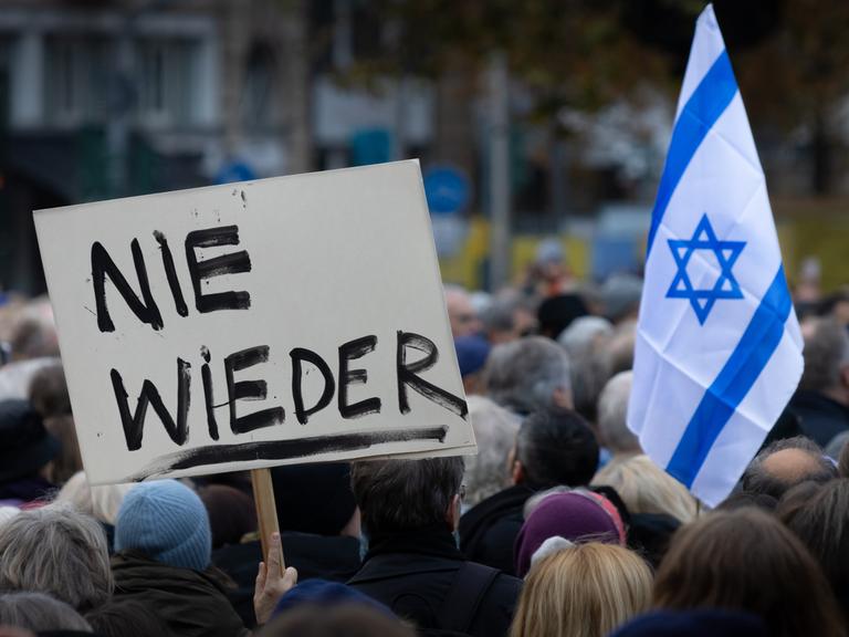 Aus einer Gruppe von Demonstrierenden ragt ein Schild mit der Aufschrift "NIE WIEDER" hervor, daneben eine israelische Flagge mit dem Davidstern.