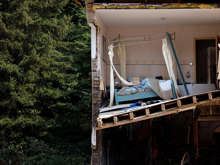 Ein zerstörtes Haus in der Eifel nach der Flutkatastrophe 2021, das Haus zeigt durchgeschnitten ein Schlafzimmer mit Bett, im Hintergrund ein Wald.