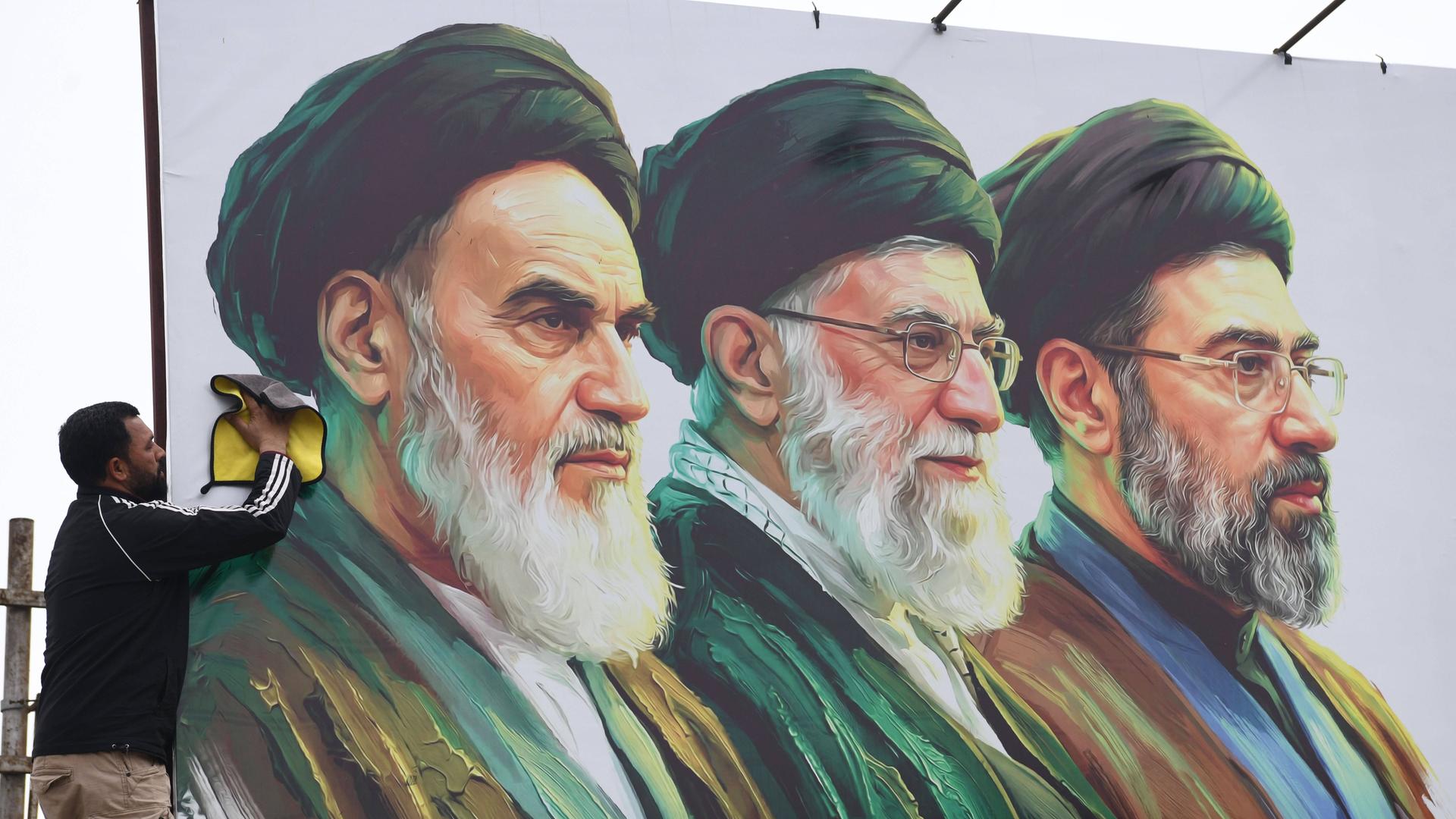Ein Mann säubert ein Plakat auf dem v.l. Ayatollah Ruhollah Chomeini, Ali Chamenei und Modschtaba Chamenei zu sehen sind. Ein Mann säubert ein Plakat auf dem v.l. Ayatollah Ruhollah Chomeini, Ali Chamenei und Modschtaba Chamenei zu sehen sind.