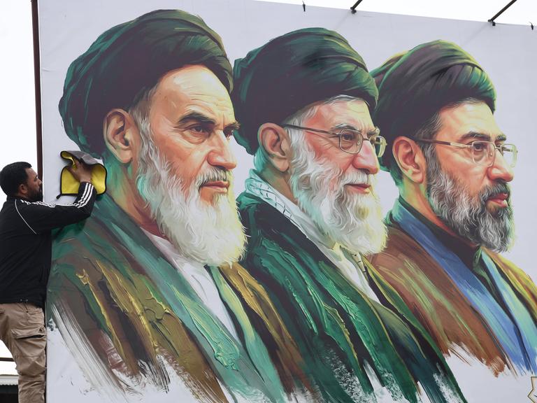 Ein Mann säubert ein Plakat auf dem v.l. Ayatollah Ruhollah Chomeini, Ali Chamenei und Modschtaba Chamenei zu sehen sind.
