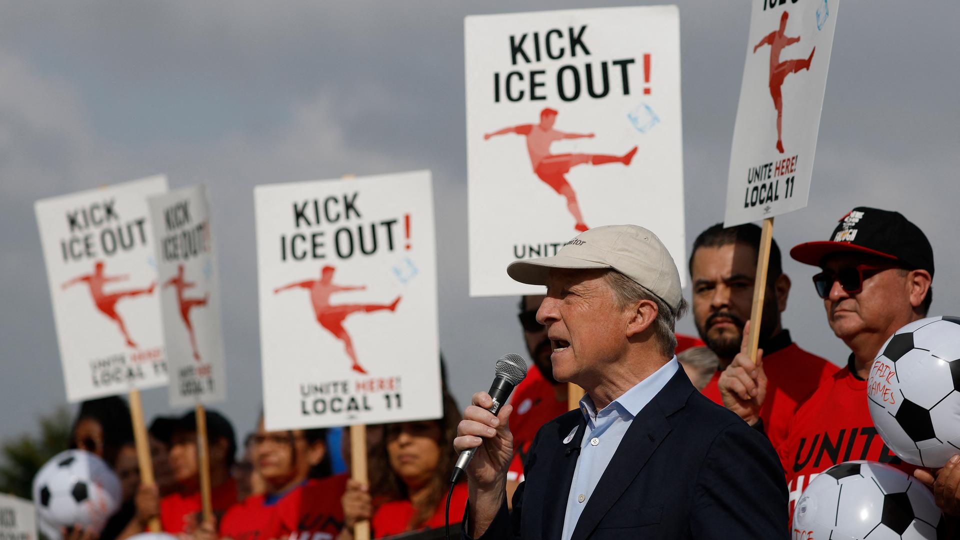 Zu sehen sind ein Redner mit Mikrofon und Protestierende in roter Kleidung. Sie halten Schilder mit der Aufschrift hoch: "Kick ICE out"