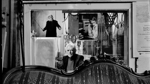 Der Komponist Johannes Fritsch sitzt hinter einem antiquierten Sofa in seinem Studio in Köln vor einer Glassscheibe, die das Studio vom Regieraum abtrennt