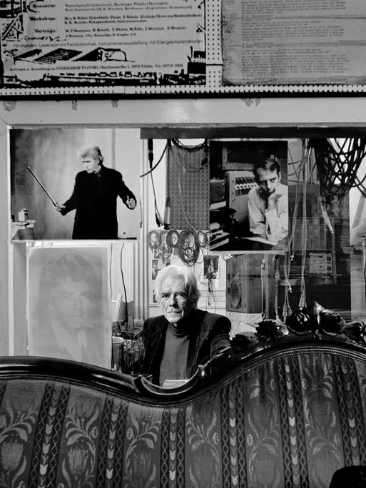 Der Komponist Johannes Fritsch sitzt hinter einem antiquierten Sofa in seinem Studio in Köln vor einer Glassscheibe, die das Studio vom Regieraum abtrennt