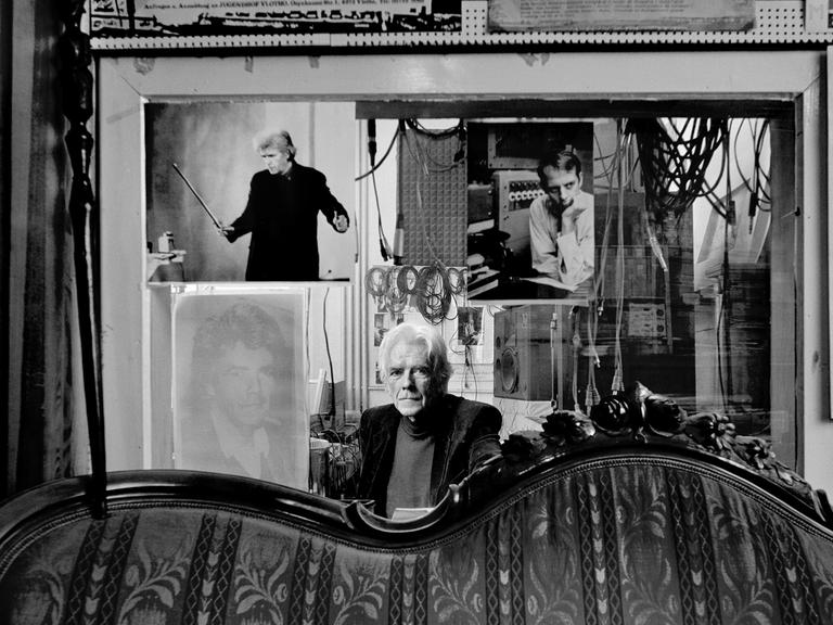 Der Komponist Johannes Fritsch sitzt hinter einem antiquierten Sofa in seinem Studio in Köln vor einer Glassscheibe, die das Studio vom Regieraum abtrennt