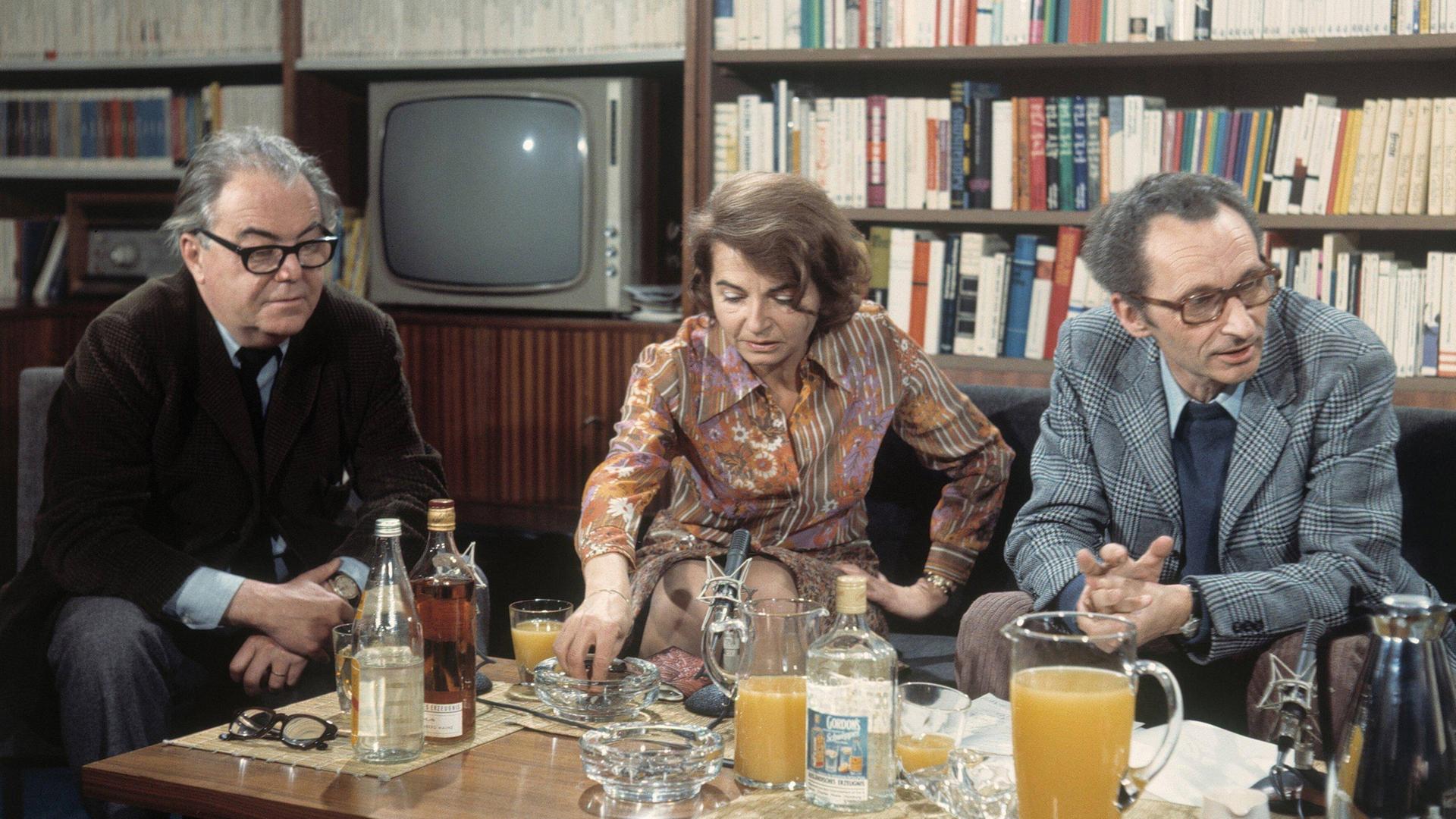 Max Frisch, Barbara König und Walter Höllerer sitzen im Jahr 1972 an einem mit Getränken beladenen Tisch vor einer Bücherwand mit Fernseher.
