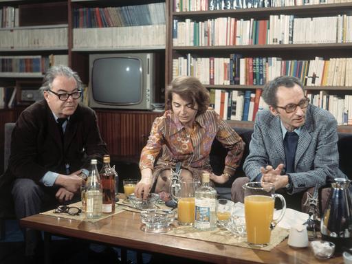 Max Frisch, Barbara König und Walter Höllerer sitzen im Jahr 1972 an einem mit Getränken beladenen Tisch vor einer Bücherwand mit Fernseher.