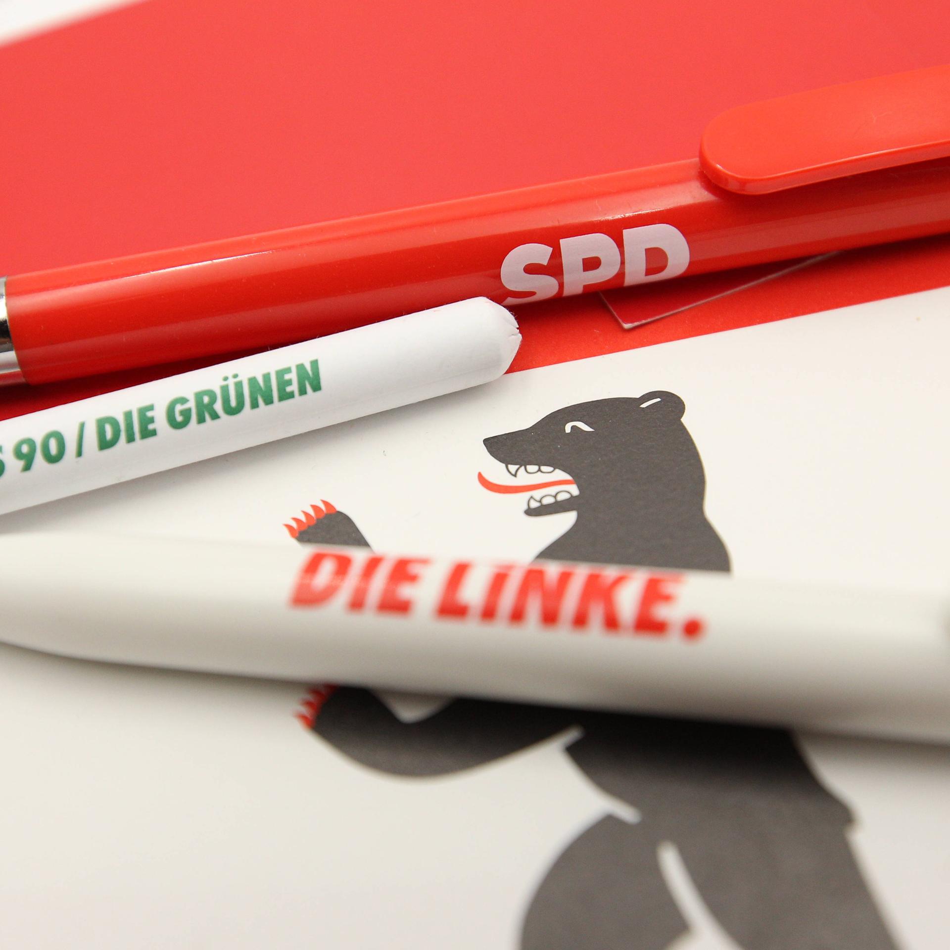 Auf einer Flagge von Berlin liegen Kugelschreiber der Parteien die Linke, SPD und Bündnis 90/Die Grünen. 