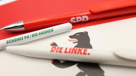 Auf einer Flagge von Berlin liegen Kugelschreiber der Parteien die Linke, SPD und Bündnis 90/Die Grünen. 