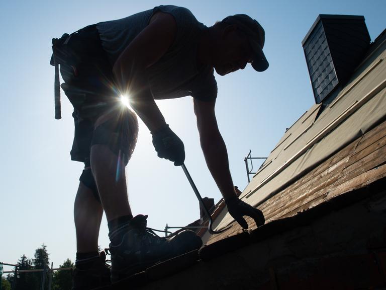 Ein Dachdecker entfernt im Gegenlicht mit einem Hammer Schindeln aus Bitumen auf einem Dach.