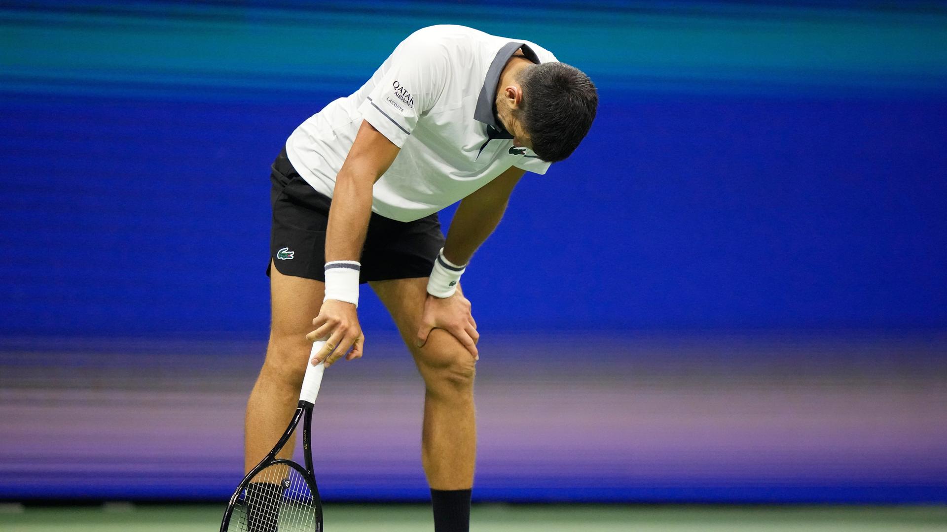 Zu sehen ist der Tennisspieler Novak Djokovic bei den US Open. Er steht auf dem Platz und schaut nach unten. Seine Haltung wirkt nachdenklich und angespannt,