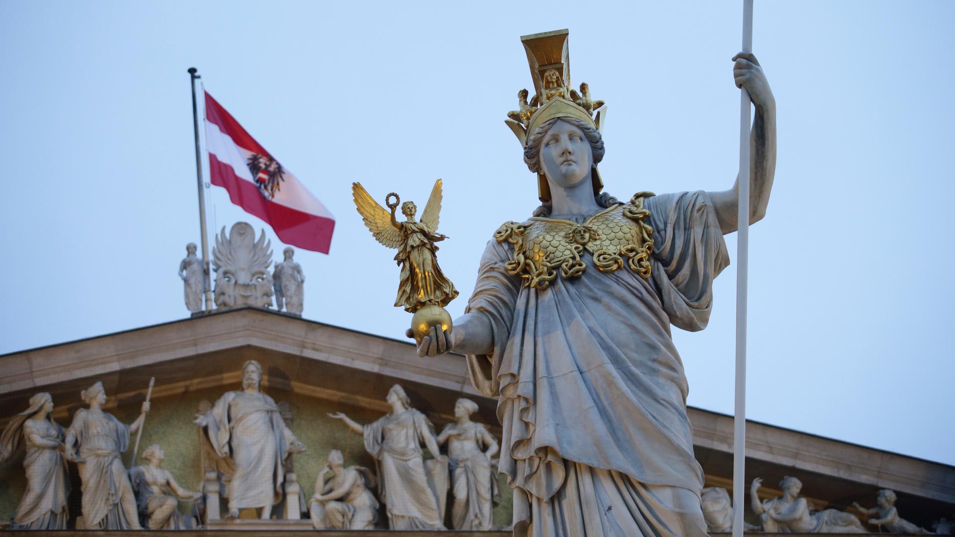 Nahaufnahme der Statue, dahinter das Dach des Parlaments mit der rot-weißen Landesflagge unter blauem Himmel. 