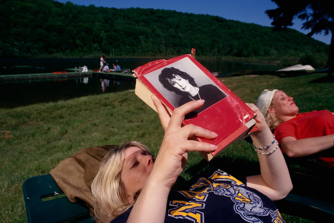 Eine Frau liegt auf einer Wiese und liest ein zerfleddertes Buch mit dem Porträt von Sue Grafton auf dem Umschlag.