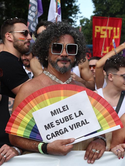 Teilnehmer der Pride-Parade 2025 in Buenos Aires. Ein Mann hält einen Fächer in Regenbogenfarben und ein Schild mit der Aufschrift "Milei me sube la carga viral".