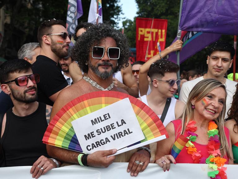 Teilnehmer der Pride-Parade 2025 in Buenos Aires. Ein Mann hält einen Fächer in Regenbogenfarben und ein Schild mit der Aufschrift "Milei me sube la carga viral".