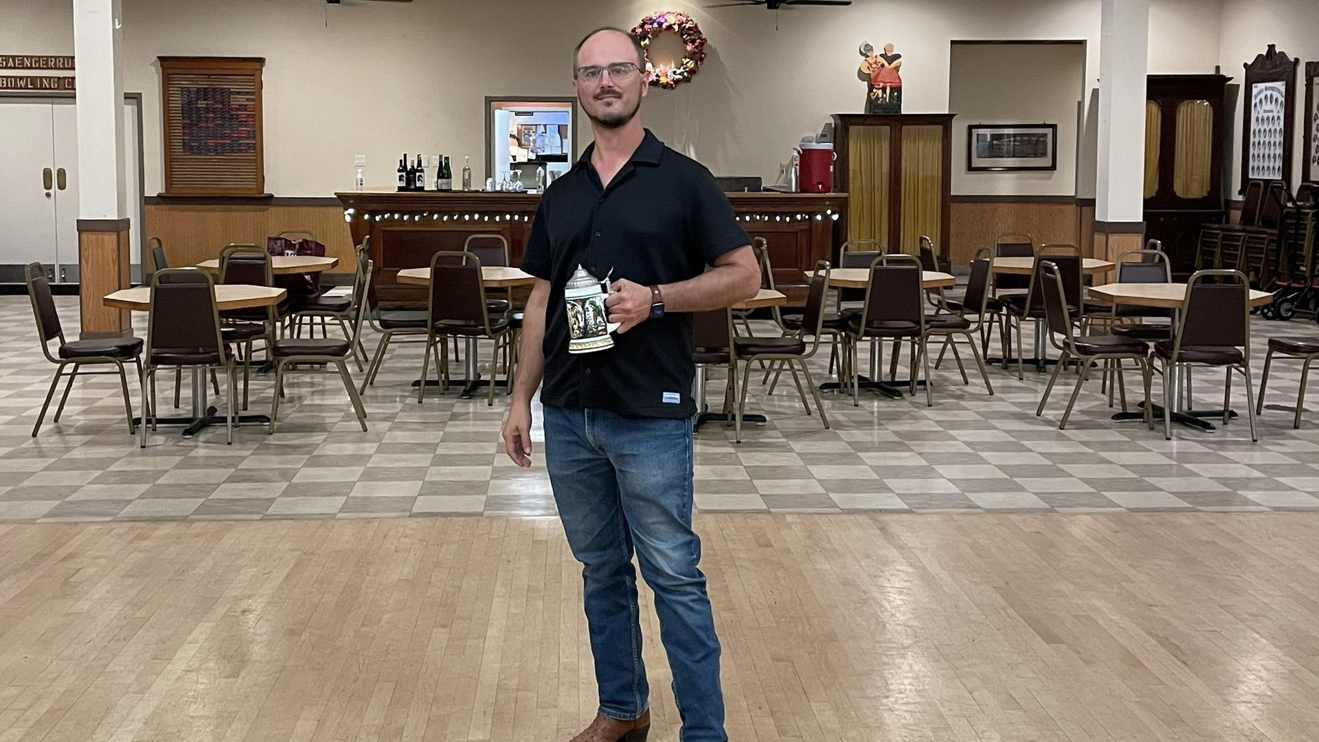 Robert Wagner steht mit einem Bierkrug in der Hand in einem großen Saal. Über ihm hängt an der Wand ein Schriftzug in altdeutscher Schrift: "Das Deutsche Lied". Robert Wagner steht mit einem Bierkrug in der Hand in einem großen Saal. Über ihm hängt an der Wand ein Schriftzug in altdeutscher Schrift: "Das Deutsche Lied".