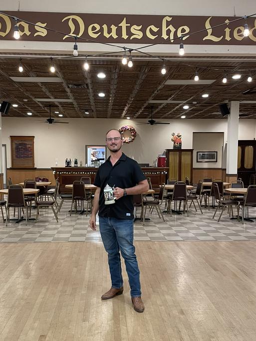 Robert Wagner steht mit einem Bierkrug in der Hand in einem großen Saal. Über ihm hängt an der Wand ein Schriftzug in altdeutscher Schrift: "Das Deutsche Lied".