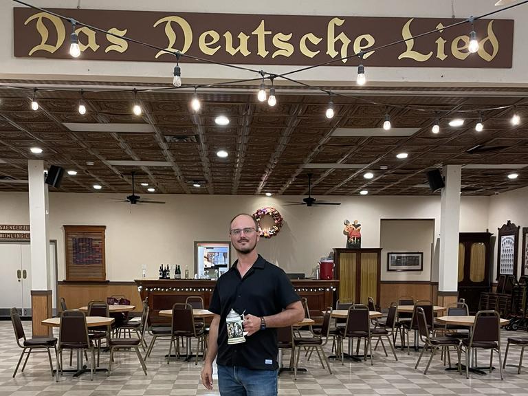 Robert Wagner steht mit einem Bierkrug in der Hand in einem großen Saal. Über ihm hängt an der Wand ein Schriftzug in altdeutscher Schrift: "Das Deutsche Lied".