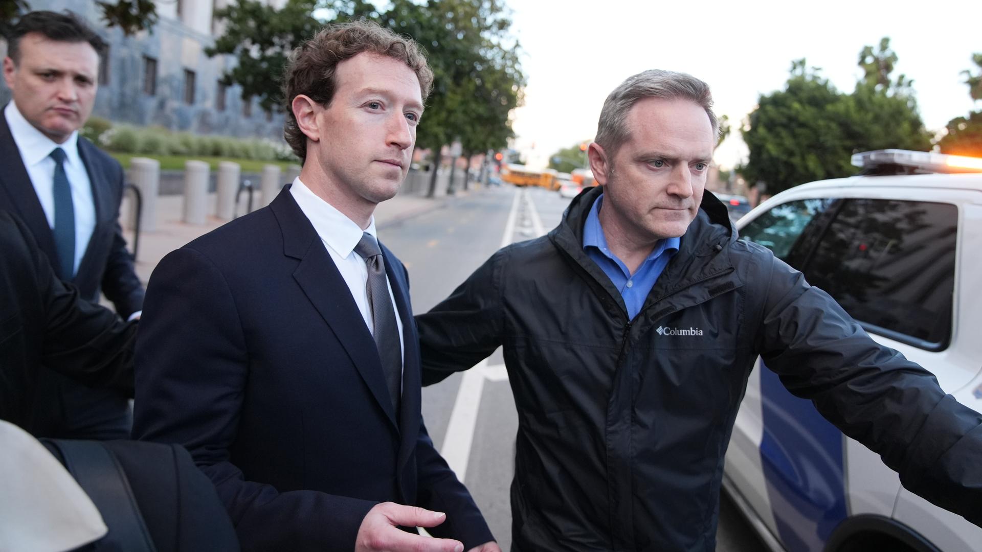 Ein Mann im dunklen Anzug verlässt begleitet von mehreren Männern ein Gerichtsgebäude und geht auf ein Fahrzeug am Straßenrand zu.