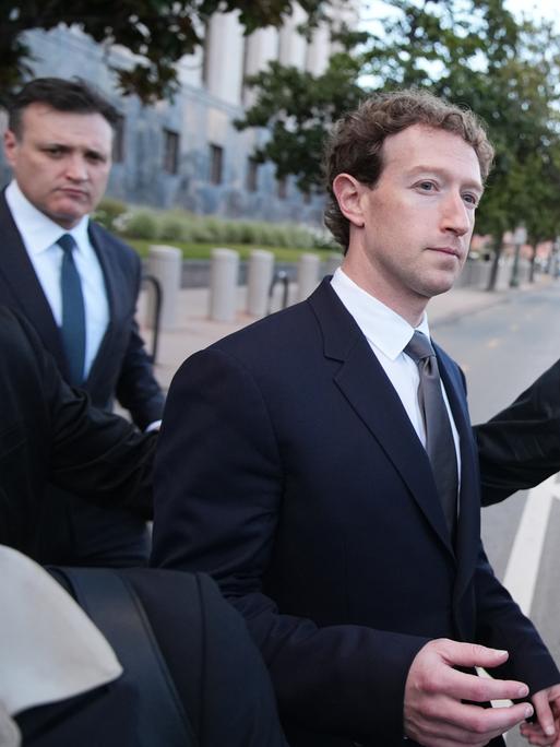 Ein Mann im dunklen Anzug verlässt begleitet von mehreren Männern ein Gerichtsgebäude und geht auf ein Fahrzeug am Straßenrand zu.