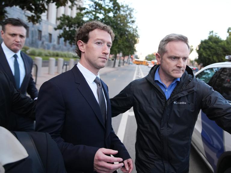 Ein Mann im dunklen Anzug verlässt begleitet von mehreren Männern ein Gerichtsgebäude und geht auf ein Fahrzeug am Straßenrand zu.