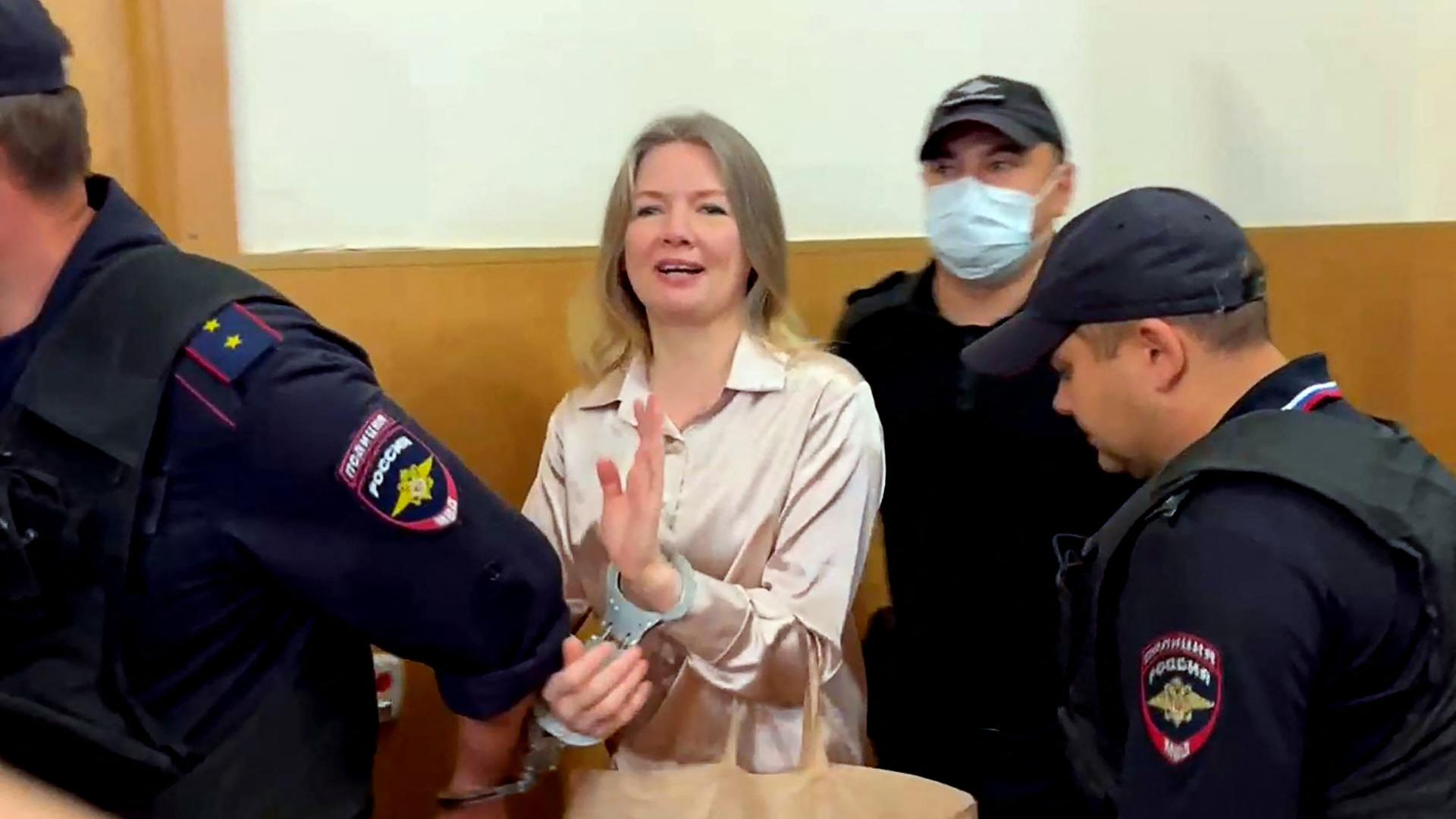 Die Journalistin Olga Komlewa wird von Beamten in den Richtersaal geführt. Sie ist mit Handschellen an einen von ihnen gefesselt und winkt.