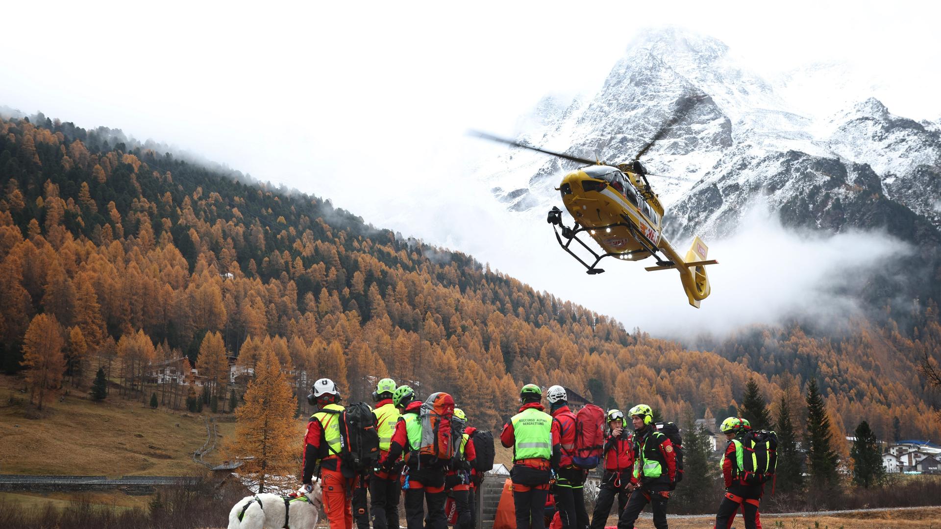 Ein Hubschrauber landet vor Bergrettern. Im Hintergrund ist ein schneebedeckter Berggipfel zu sehen und ein kleinerer Berg mit Fichtenwald.