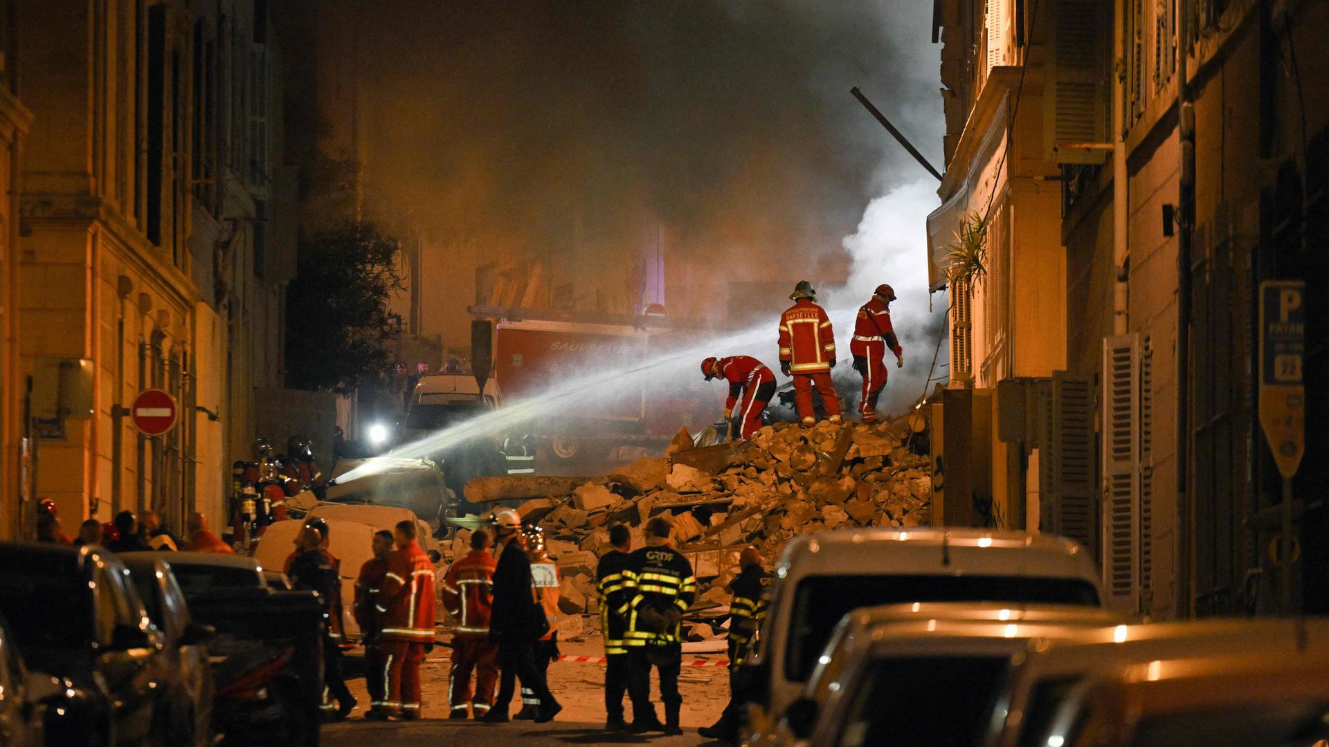 Rettungskräfte arbeiten an der Stelle eines eingestürzten Gebäudes in der südfranzösischen Hafenstadt Marseille. Zu sehen ist ein Trümmerhaufen, auf dem Feuerwehrleute stehen. Aus einem Feuerwehrschlauch kommt Wasser.