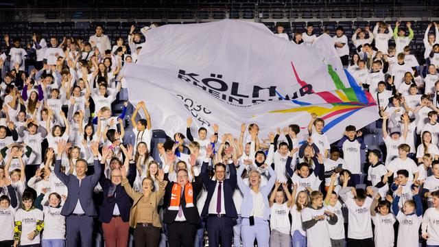 Viele Sportler und Politiker sind in Köln versammelt. Sie jubeln und reißen die Arme in die Luft. Im Hintergrund ist ein Plakat zu sehen auf dem steht "KölnRheinRuhr".