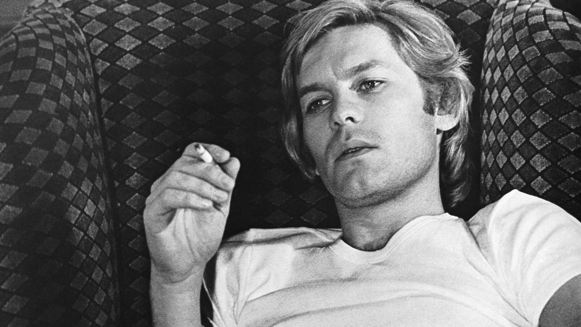 Das Foto zeigt eine schwarz-weiß Aufnahme des Schauspielers Helmut Berger. Er sitzt in einem Sessel und raucht eine Zigarrette.