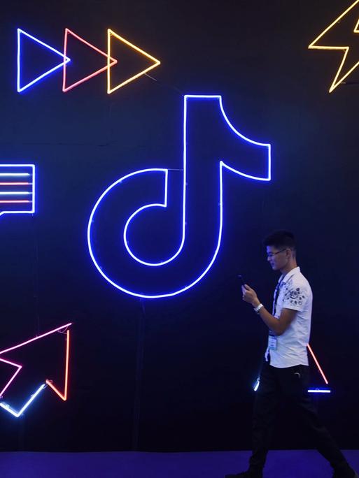 Ein Mann läuft mit seinem Smartphone vor einer Wand entlang, an der in Neonfarben das TikTok-Logo, Herzen und Pfeile prangen.