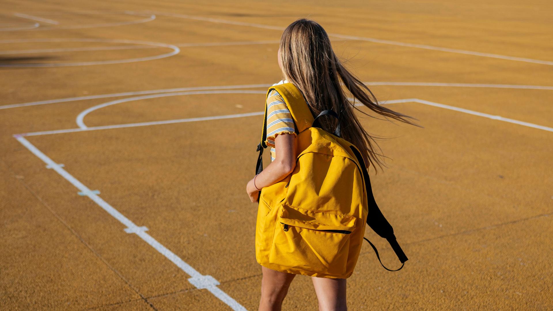 Ein junges Mädchen trägt einen Rucksack und geht über einen Basketballplatz.