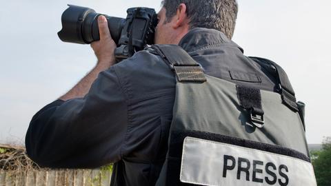 Blick auf einen Fotografen, der mit dem Rücken zum Betrachter steht. Er macht gerade mit seinen Apparat eine Aufnahme und trägt eine Sicherheitsweste mit der Aufschrift "Press". 